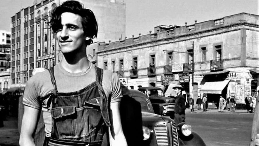 Imagen en blanco y negro de Roberto Cobo, actor con peto vaquero, sonriendo ligeramente. Fondo urbano con edificios y vehículos antiguos.