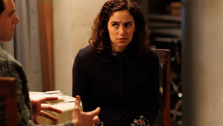 Dos personas, una mujer con chaqueta oscura y un hombre con camisa estampada, mantienen una conversación tensa sentados a una mesa con mantel blanco.