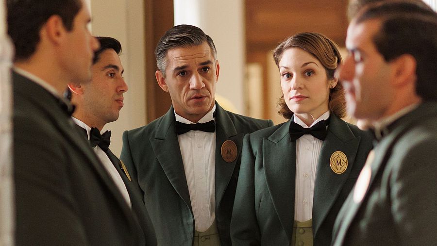 Cuatro empleados de "La Moderna", con uniformes verdes y pines dorados con una "M", en una escena que parece una bienvenida a una nueva empleada.