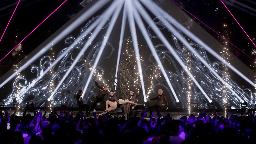 Actuación de Melody en el escenario con bailarines, vestuario claro y oscuro, iluminación impactante y efectos especiales. Público grabando con móviles.