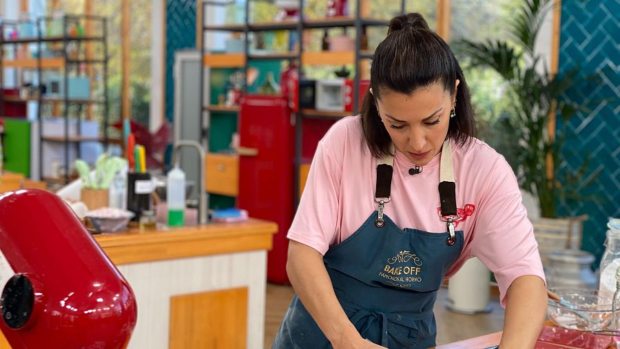 Presentadora con delantal azul marino amasando en una cocina con azulejos turquesa y utensilios de repostería.  Cabello recogido.