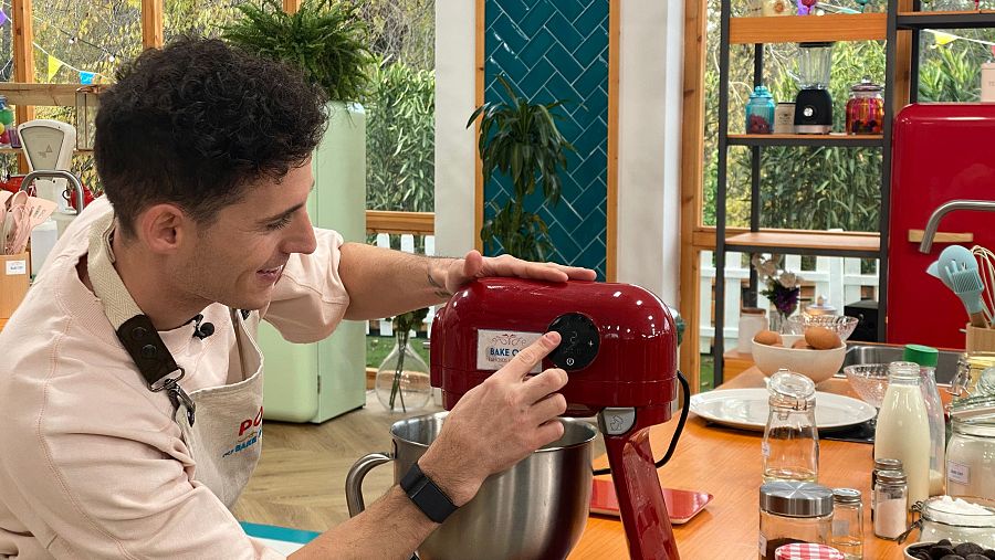 Pol Espargaró en una cocina, con delantal, ajusta una batidora roja rodeado de ingredientes para cocinar.
