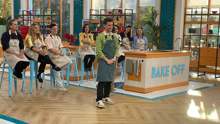 Mario Marzo en el programa "Bake Off", con delantal verde, sonríe a cámara.  Detrás, otros concursantes con delantales similares.  Ambiente de estudio de televisión.