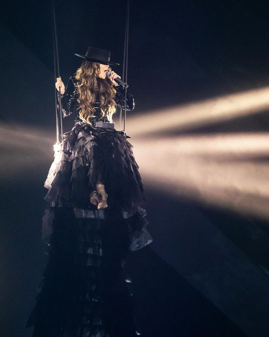 Cantante en un columpio a gran altura, con vestido y sombrero negros, sujetando un micrófono durante una actuación con iluminación dramática.