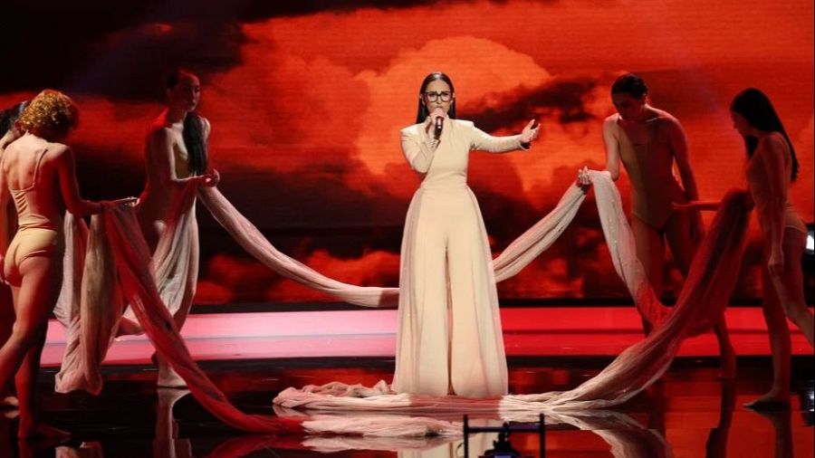 Cantante con traje beige y gafas actuando en escenario rojo con bailarinas y telas, creando una atmósfera dramática.