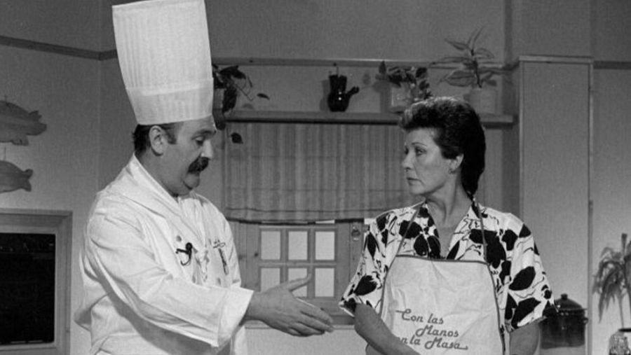 Escena de cocina en blanco y negro: un chef instruye a una presentadora con delantal. Decoración con siluetas de peces.