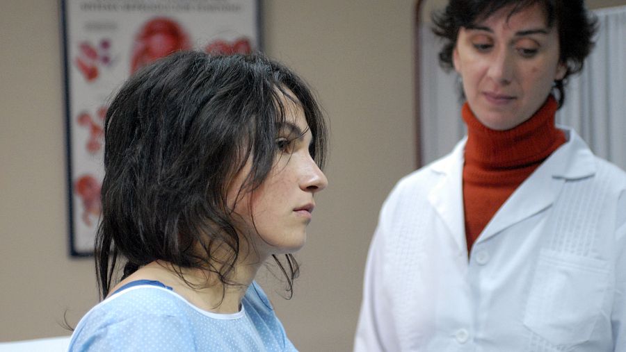 Dos mujeres, una paciente con bata azul y una doctora con bata blanca y jersey naranja, se miran con seriedad en una consulta médica. Póster anatómico en la pared.