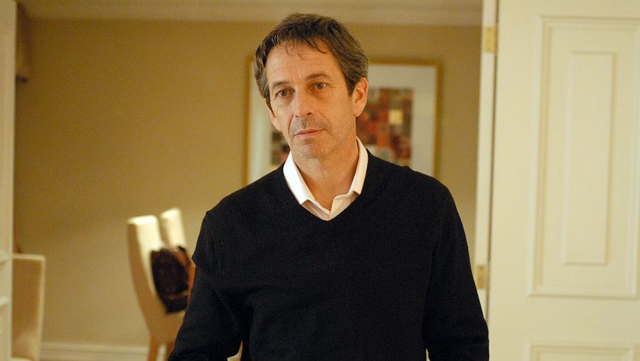 Escena interior de la película "La buena vida" (2008) con Alfredo Castro, quien viste jersey negro sobre camisa blanca. Iluminación cálida.