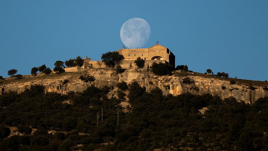 Ermita de color ocre con torre, situada en colina rocosa bajo una gran luna llena.  Atmósfera serena y solitaria al amanecer o anochecer.