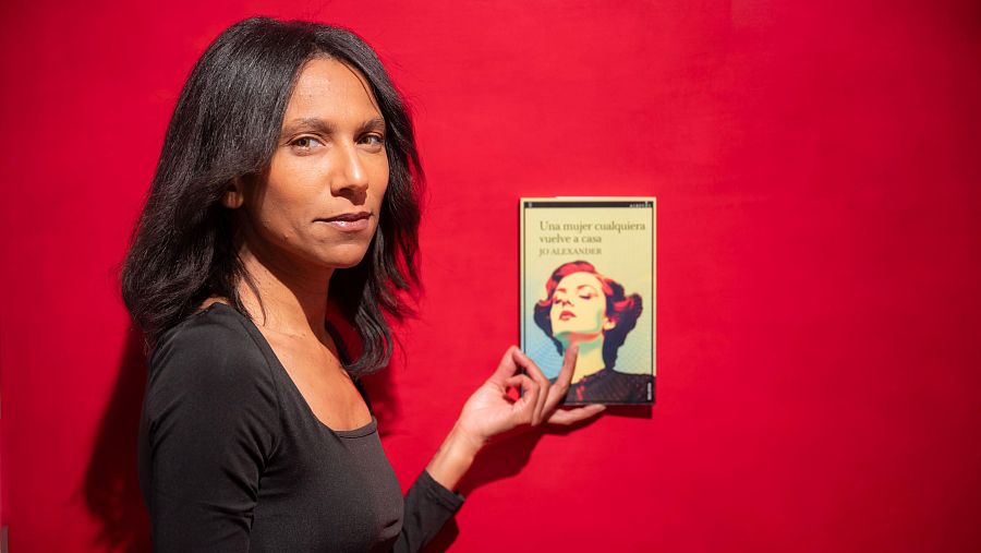 Joana Sánchez (Jo Alexander) presenta su libro "Una dona quasevol torna a casa".  Cabello largo y oscuro, camiseta negra, fondo rojo vibrante.