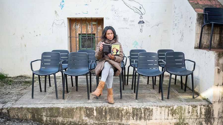 Mujer leyendo un libro con cubierta amarilla en silla de plástico, en espacio exterior con suelo embaldosado y pared con grafitis.