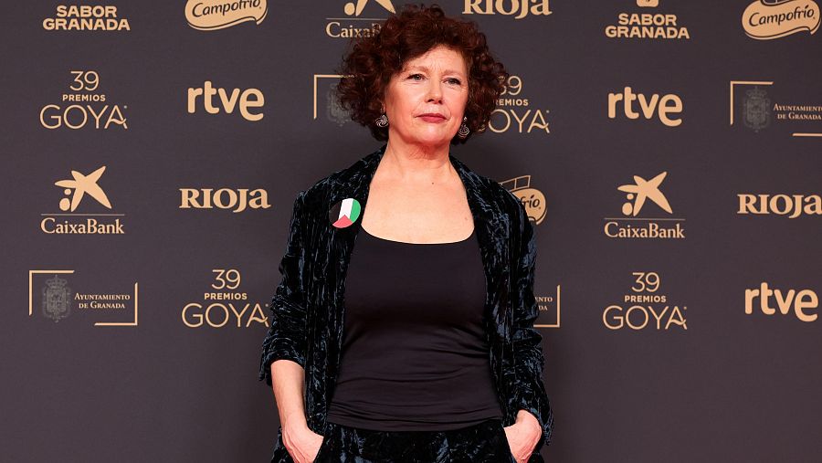 Icíar Bollaín posa en un photocall de los Premios Goya 2025, con traje oscuro y un pin.  Fondo negro con logos de patrocinadores.