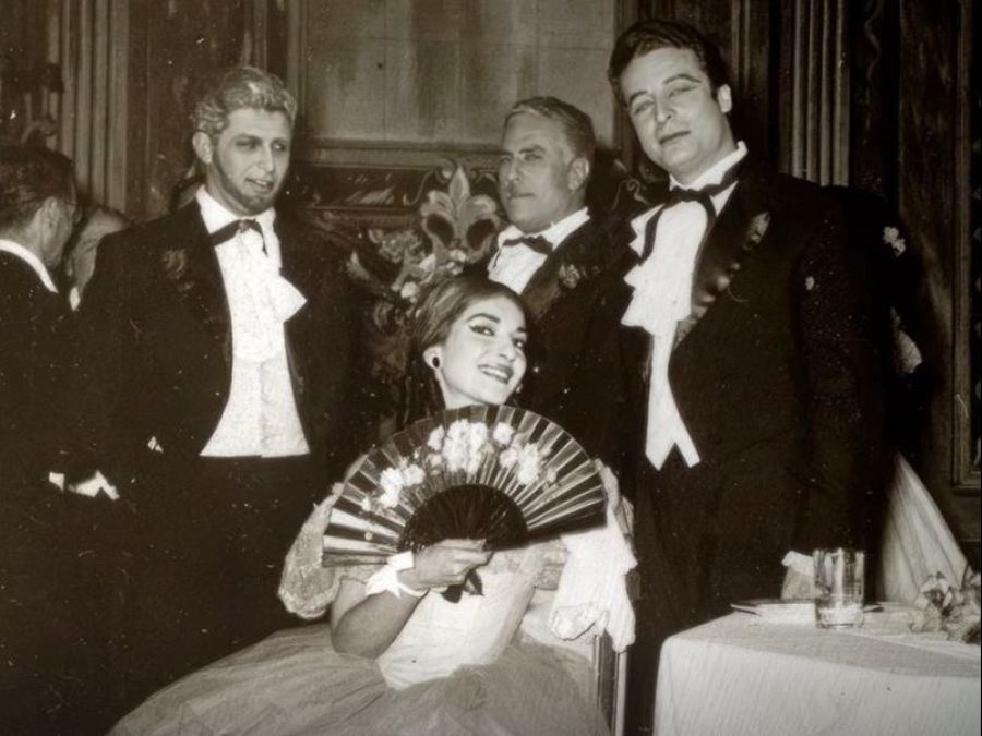 Retrato en blanco y negro de María Callas, sentada con un abanico, junto a tres hombres en traje oscuro. Fondo oscuro y mesa con mantel blanco.