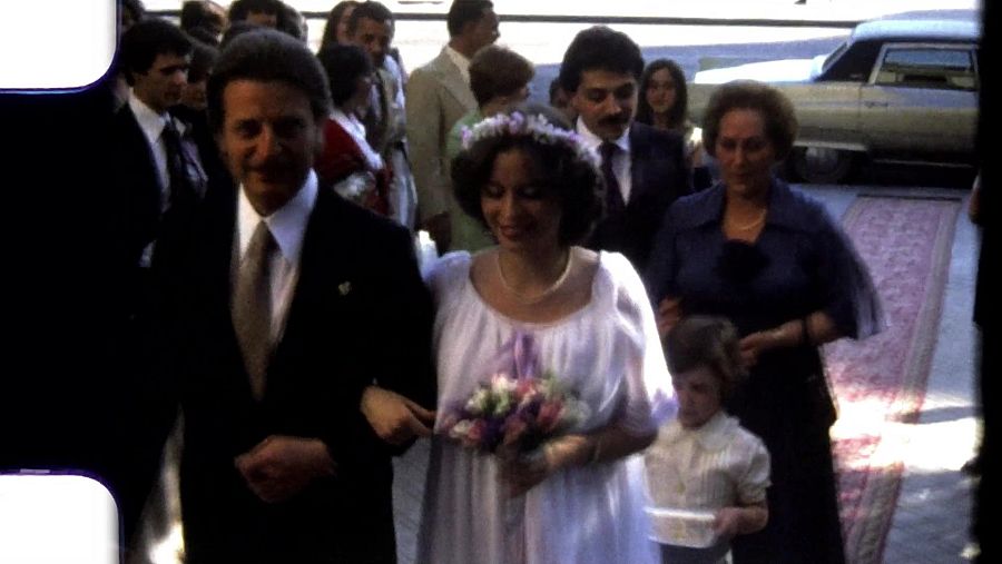 Boda al aire libre. Pareja vestida de gala camina por alfombra roja, precedida por una niña.  Invitados y un Cadillac clásico de fondo.