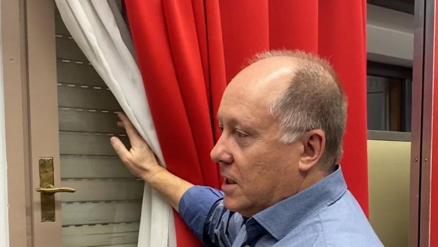 Hombre con camisa azul observa por una ventana con persianas, apartando cortinas blancas y rojas en un estudio de grabación.