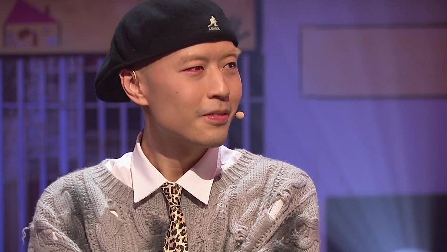 Rapero con gorro Kangol, jersey gris y corbata de leopardo, usando micrófono de solapa en un plató de televisión con fondo morado.