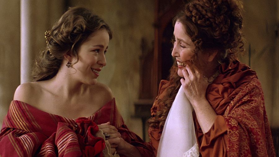 Dos actrices en escena de época, vestidas con atuendos antiguos, interactúan con cercanía y complicidad. Una de ellas parece arreglar el vestuario o maquillaje de la otra.