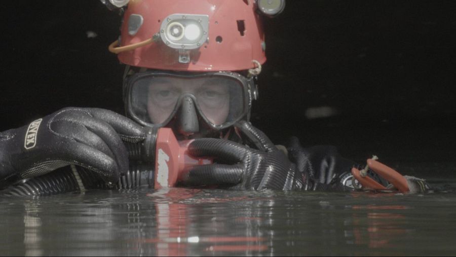 Buzo con casco rojo y guantes realiza un rescate exitoso en aguas oscuras, sosteniendo un objeto rectangular rojo.  Imagen de un documental.