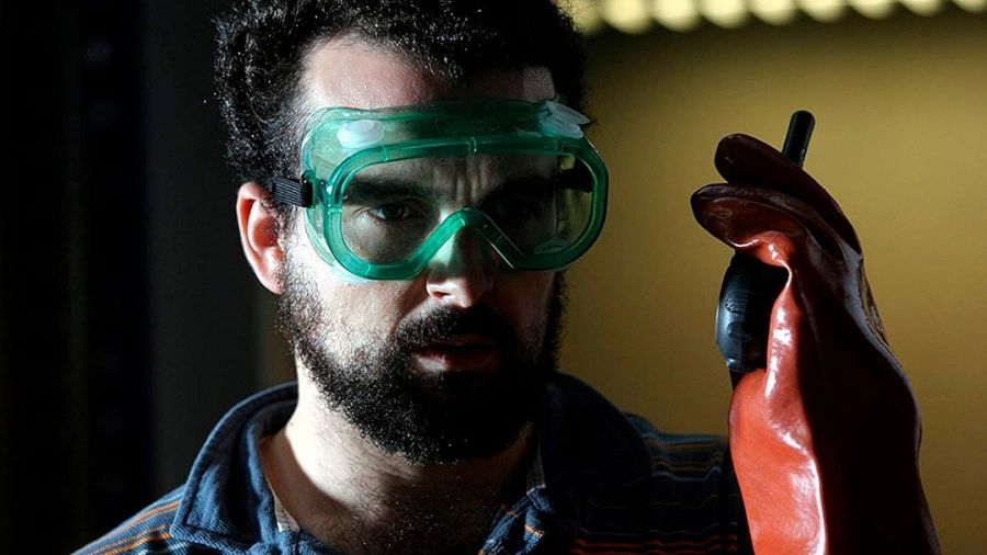 Nacho Vigalondo, con gafas protectoras verdes y guante naranja, concentrado en un set de rodaje con iluminación tenue y fondo desenfocado.
