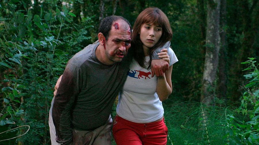 Escena de 'Los cronocrímenes' con Karra Elejalde herido (sangre en cara y ropa) apoyándose en Bárbara Goenaga en un bosque.