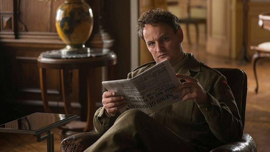 Hombre en uniforme militar leyendo un periódico, "The News Guardian", sentado en un sillón de cuero en una habitación con muebles oscuros.  Ambiente tranquilo.