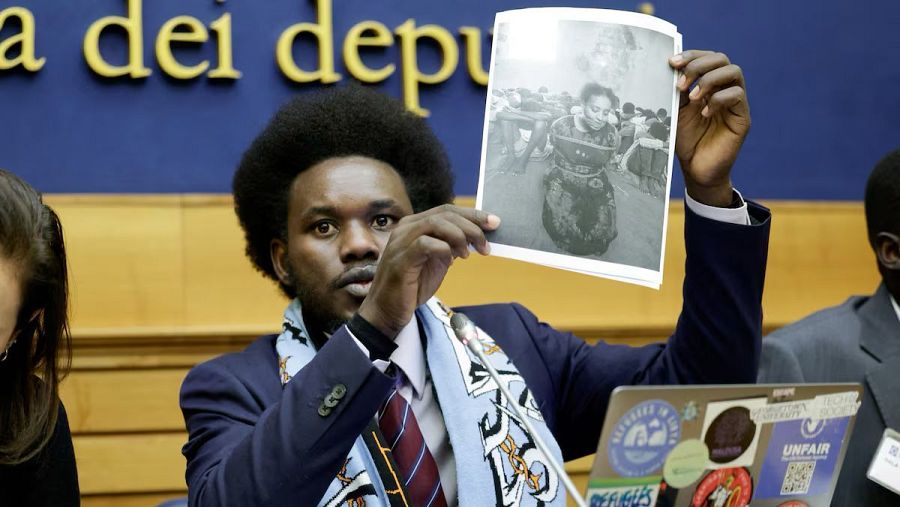 David Yambio muestra una foto de una niña;  su portátil tiene pegatinas de 'Refugees in Libya' y 'Unfair'; el fondo parece una sala de conferencias.