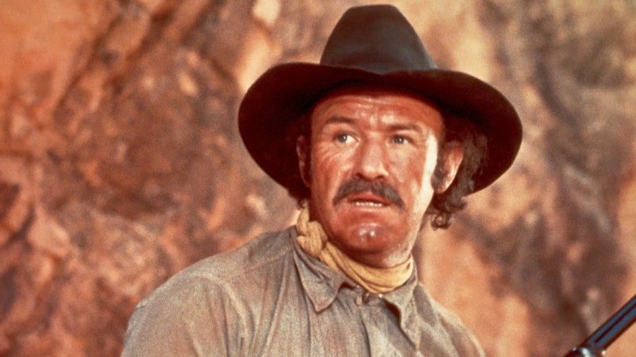 Imagen de Gene Hackman en una escena de película; viste sombrero de vaquero, camisa clara y pañuelo. Expresión seria, fondo desértico borroso.  Se ve parte de un rifle.