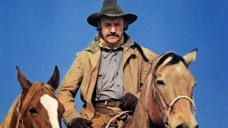 Gene Hackman monta un caballo marrón claro en una escena de 'Muerde la bala'. Lleva sombrero, chaqueta beige y camisa azul. Se ve parte de otro caballo.