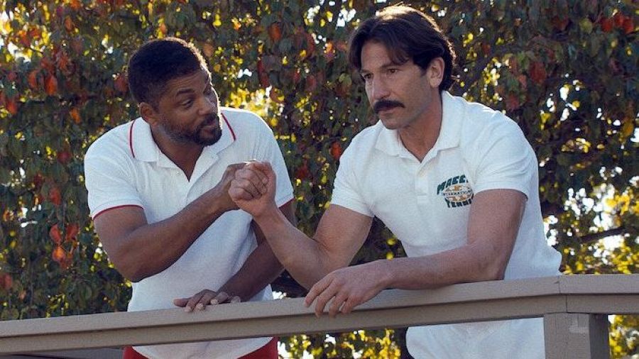Escena de película: dos hombres con polos blancos se dan la mano en una barandilla, fondo con árboles.  Uno viste un polo con ribetes rojos; el otro, un polo con el logo "Macci International Tennis".