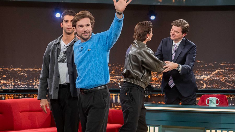 Tres actores invitados en un programa de televisión. Uno de ellos saluda mientras el presentador, de traje, estrecha la mano de otro invitado. Se ve un sofá y una mesa con una taza con el logo del programa.