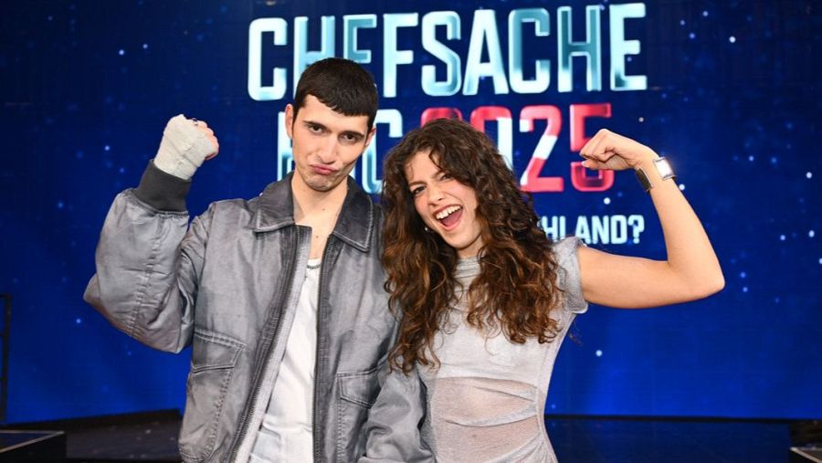 Dos personas, un hombre con chaqueta gris y una mano vendada y una mujer con camiseta gris clara, celebran con los brazos levantados.  Fondo con el logo "CHEFSACHE" y el año 2025.