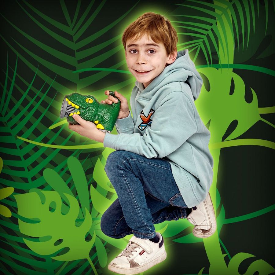 Niño pelirrojo con sudadera verde claro y vaqueros sostiene un dinosaurio pompero verde oscuro, sonriendo a cámara sobre fondo tropical.