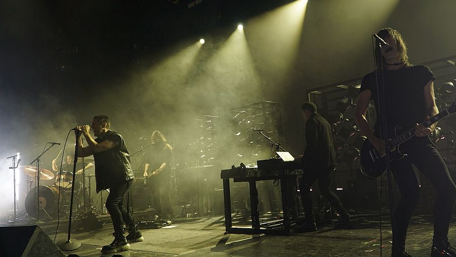 Actuación en directo de una banda de rock en un escenario oscuro y con humo, con iluminación tenue y cinco músicos vestidos de negro.