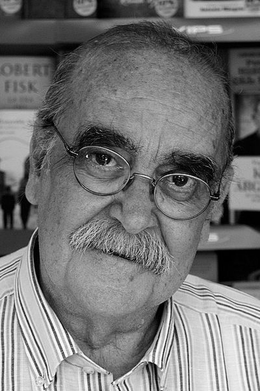 Primer plano de José Antonio Labordeta con gafas y camisa a rayas en un entorno con libros. Imagen en blanco y negro.