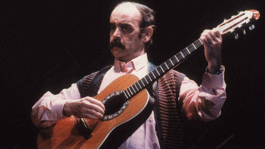 Cantautor con camisa a rayas rosas y chaleco oscuro tocando una guitarra acústica en un escenario con fondo oscuro.