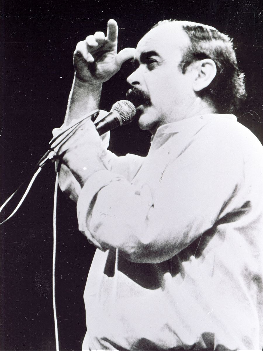 Imagen en blanco y negro de José Antonio Labordeta en un escenario, con micrófono en mano, gesticulando mientras parece cantar o hablar.