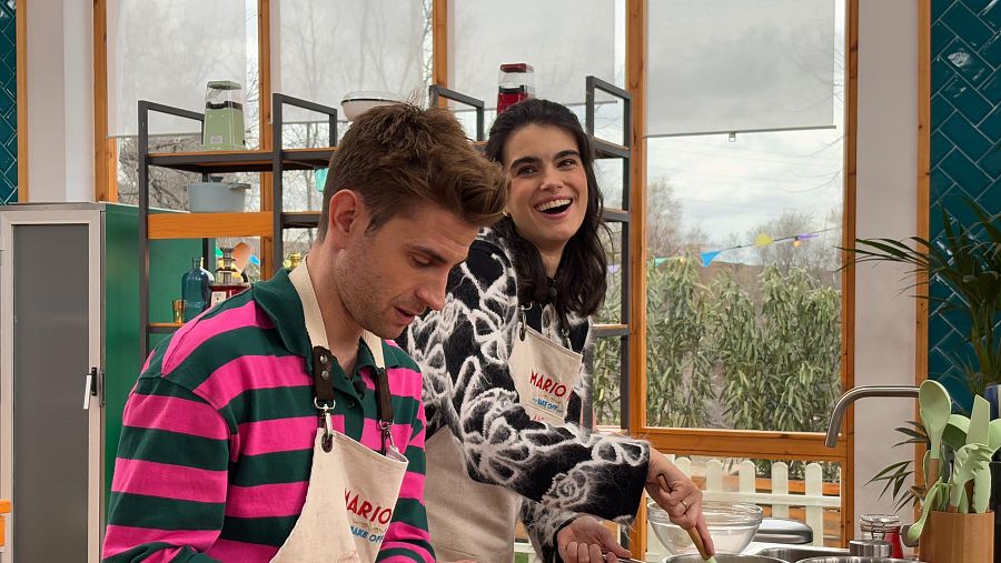 Escena de 'Bake Off: famosos al horno' mostrando a dos concursantes, uno concentrado en su preparación y otro sonriendo mientras remueve un bol con una espátula de madera en una cocina equipada.