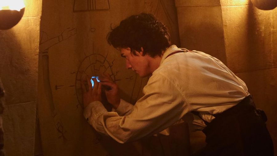 Joven con camisa clara y pantalones oscuros examina grabados en una pared de piedra oscura, activando una luz azul al tocar un círculo.