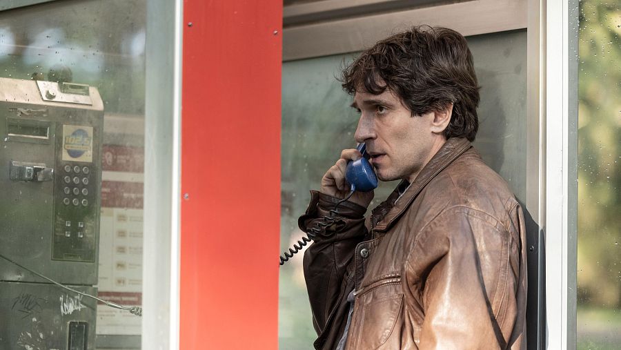 Hombre concentrado hablando por teléfono en una deteriorada cabina telefónica antigua con teclado numérico y auricular azul.