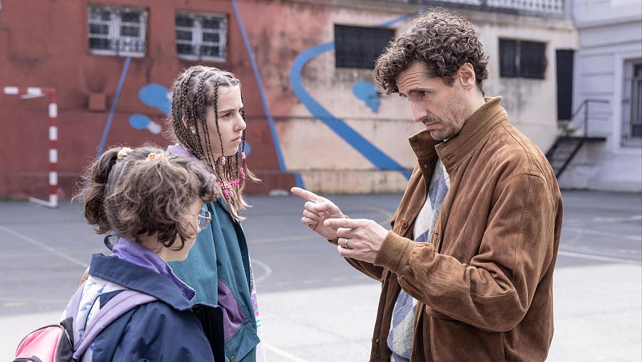 Andoni Agirregomezkorta conversa con dos jóvenes en un patio escolar.  Una lleva mochila rosa y chaqueta azul, la otra una chaqueta verde.  Fondo con muro y grafitis.