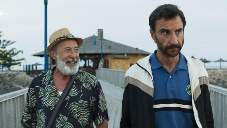 Dos hombres conversan mientras caminan por un muelle. Uno lleva sombrero de paja y camisa estampada; el otro, chaqueta deportiva. Al fondo, se ve el mar y una construcción.