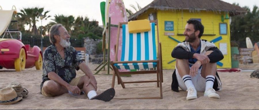Dos hombres, uno mayor y otro joven, sentados en una playa arenosa, conversan junto a una tumbona y una caseta con tablas de surf.  Se observan detalles de su ropa.
