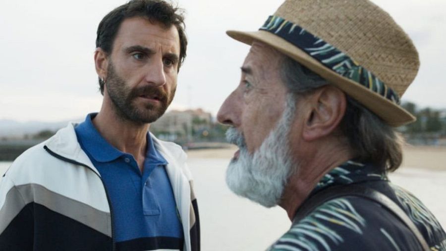 Dos hombres, uno joven con chaqueta deportiva y otro mayor con sombrero de paja, conversan seriamente en una playa con edificios al fondo.  Ambiente tenso.