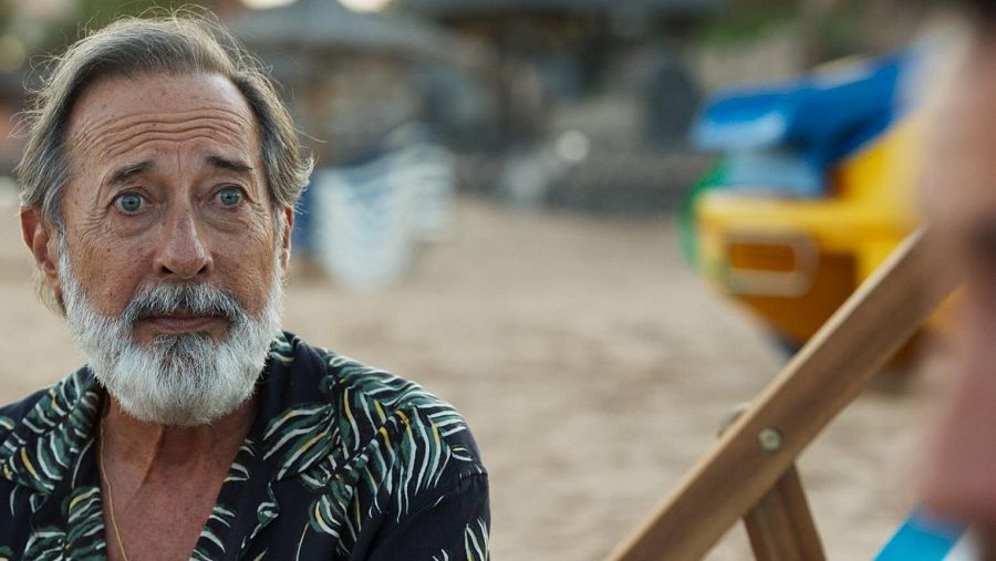 Primer plano de Juan Diego en 'Playa de Lobos'.  Camisa oscura, expresión seria, playa de fondo con embarcación y sombrilla.