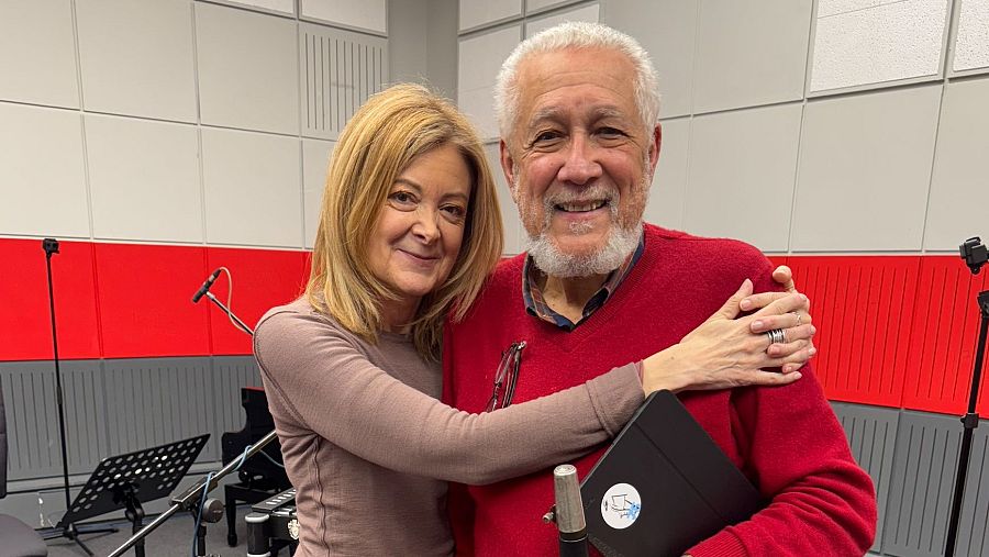 Imagen de José Sacristán y una mujer sonriendo en un estudio de grabación. Él lleva un jersey rojo y ella uno marrón claro; él sostiene una carpeta negra. Ambiente cálido y distendido.