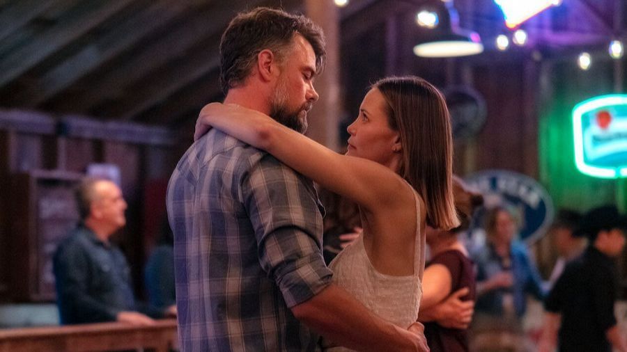 Pareja baila abrazada en ambiente rústico y festivo, con iluminación tenue. Él viste camisa de cuadros; ella, top de tirantes. Fondo con gente bailando y mesas.