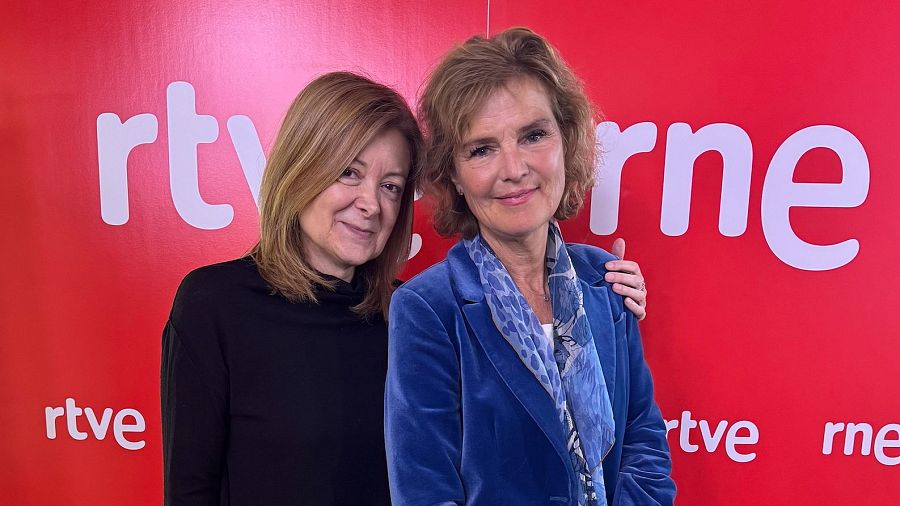 Dos mujeres, una con jersey negro y otra con chaqueta azul, posan ante un fondo rojo con el logo de RTVE.  Miran a la cámara.