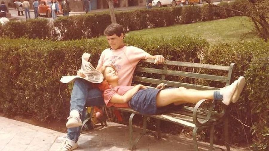 Pareja en banco de parque: él lee periódico, ella descansa.  Ambiente tranquilo, césped y arbustos visibles.