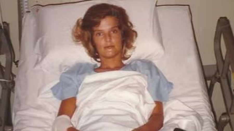 Mujer en cama de hospital con bata azul, pelo rojizo y una mano vendada. Expresión seria.