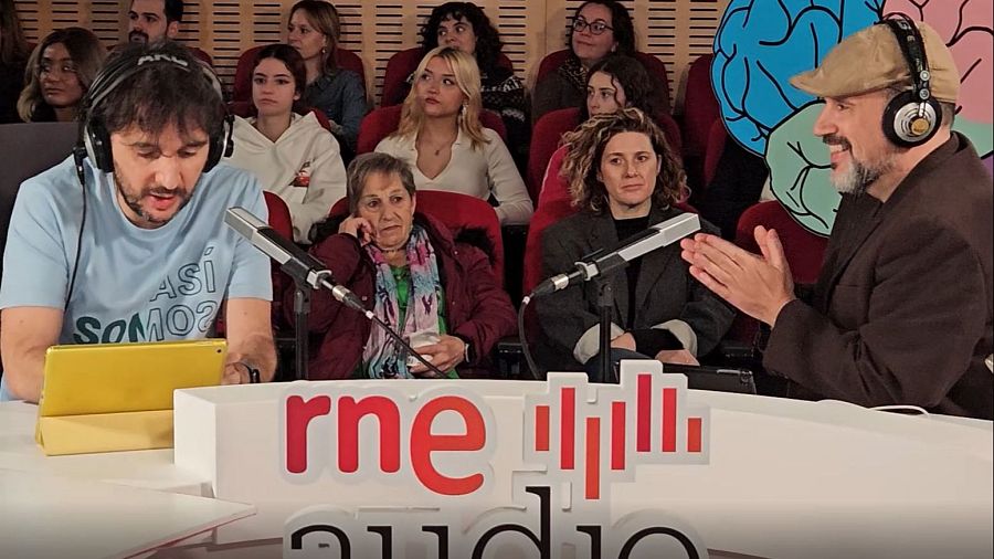Dos presentadores graban un podcast en estudio, uno con camiseta azul y tablet, el otro con boina beige. Público presente en butacas rojas.